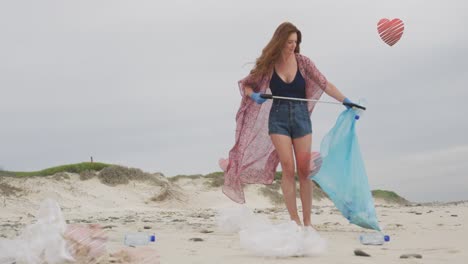 Animation-of-hearts-over-caucasian-woman-picking-up-rubbish-on-beach