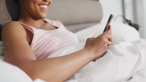 Happy-african-american-woman-laying-in-bed,-using-smartphone-in-bedroom