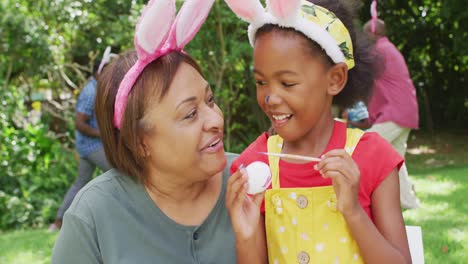 Animation-of-happy-african-american-grandmother-and-granddaughter-painting-easter-eggs-in-garden