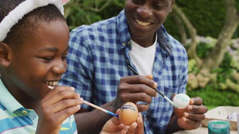 Animation-of-happy-african-american-father-and-son-painting-easter-eggs-together-in-garden