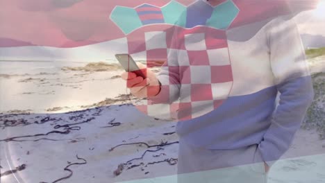 Animación-De-La-Bandera-De-Croacia-Sobre-Un-Hombre-Caucásico-Mayor-Usando-Un-Teléfono-Inteligente-En-La-Playa
