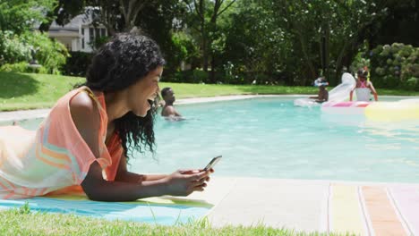 Happy-african-american-woman-relaxing-at-pool-with-family,-using-smartphone