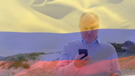 Animation-Der-Flagge-Von-Kolumbien-über-Einem-älteren-Kaukasischen-Mann,-Der-Sein-Smartphone-Am-Strand-Benutzt