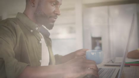 Animation-of-lights-over-focused-african-american-man-drinking-coffee-and-working-on-laptop
