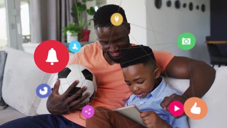 Animation-Von-Symbolen-über-Einem-Glücklichen-Afroamerikanischen-Vater-Und-Sohn,-Die-Sich-Ein-Fußballspiel-Auf-Dem-Smartphone-Ansehen