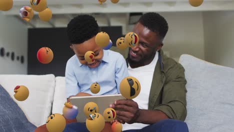 Animation-of-emoticons-over-happy-african-american-father-and-son-using-tablet-on-sofa