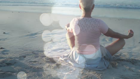 Animation-of-light-spots-over-senior-caucasian-woman-practicing-yoga-at-beach-and-meditating