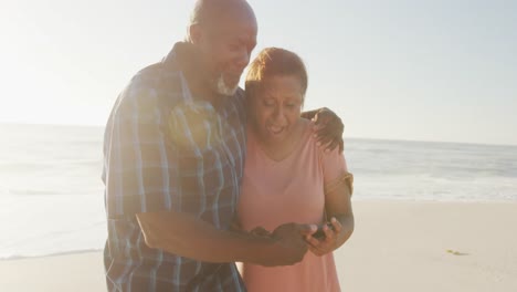 Älteres-Afroamerikanisches-Paar-Hält-Händchen-Und-Benutzt-Smartphone-Am-Sonnigen-Strand