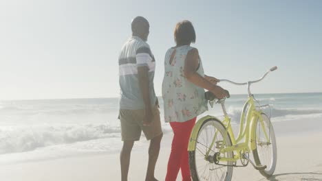 Älteres-Afroamerikanisches-Paar,-Das-Mit-Dem-Fahrrad-Am-Sonnigen-Strand-Spaziert