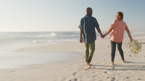 Smiling-senior-african-american-couple-holding-hands-and-walking-on-sunny-beach