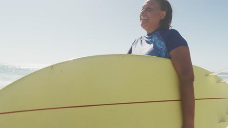 Glückliche-ältere-Afroamerikanische-Frau,-Die-Mit-Surfbrett-Am-Sonnigen-Strand-Spaziert