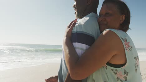 Lächelndes-älteres-Afroamerikanisches-Paar,-Das-Sich-Am-Sonnigen-Strand-Umarmt-Und-Auf-Das-Meer-Blickt