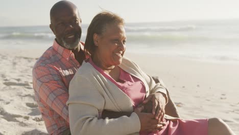 Smiling-senior-african-american-couple-embracing-and-sitting-on-sunny-beach