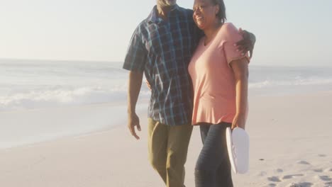 Smiling-senior-african-american-couple-embracing-and-walking-on-sunny-beach