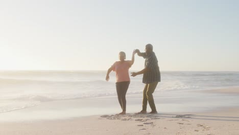 Smiling-senior-african-american-couple-holding-hands-and-dancing-on-sunny-beach