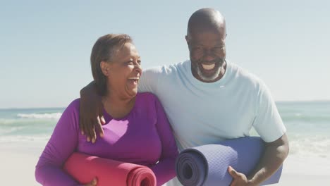 Porträt-Eines-Glücklichen-älteren-Afroamerikanischen-Paares,-Das-Yogamatten-Am-Sonnigen-Strand-Hält