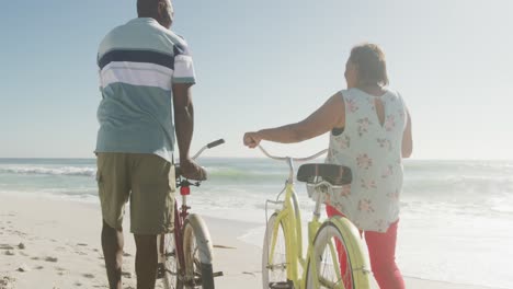 Smiling-senior-african-american-couple-walking-with-bicycles-on-sunny-beach