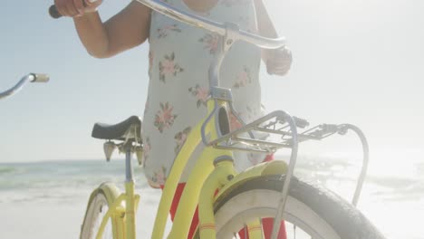 Lächelndes-älteres-Afroamerikanisches-Paar,-Das-Mit-Fahrrädern-Am-Sonnigen-Strand-Spaziert