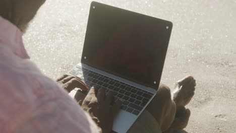 Senior-african-american-man-sitting-and-using-laptop-on-sunny-beach