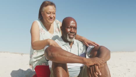 Lächelndes-älteres-Afroamerikanisches-Paar,-Das-Sich-Am-Sonnigen-Strand-Umarmt-Und-Auf-Das-Meer-Blickt