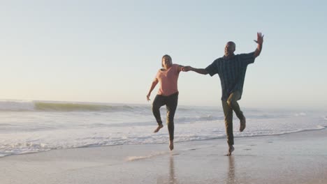 Smiling-senior-african-american-couple-holding-hands-and-running-on-sunny-beach