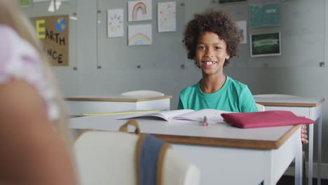 Video-of-happy-biracial-boy-sitting-at-desk-in-classsroom