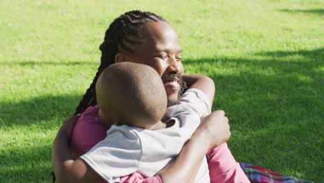 Video-Eines-Glücklichen-Afroamerikanischen-Vaters-Und-Sohnes,-Die-Im-Gras-Picknicken-Und-Sich-Umarmen