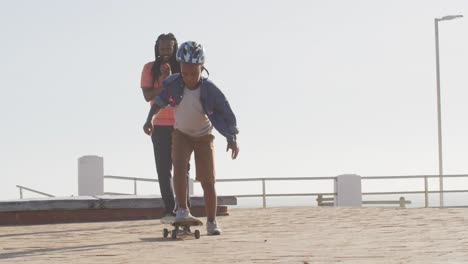 Video-Eines-Glücklichen-Afroamerikanischen-Vaters,-Der-Seinem-Sohn-Das-Skateboarden-Auf-Der-Promenade-Beibringt