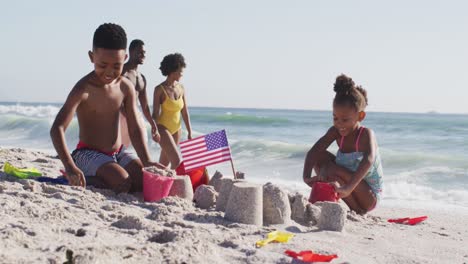 Familia-Afroamericana-Sonriente-Construyendo-Castillos-De-Arena-Con-Bandera-Americana-En-La-Playa-Soleada
