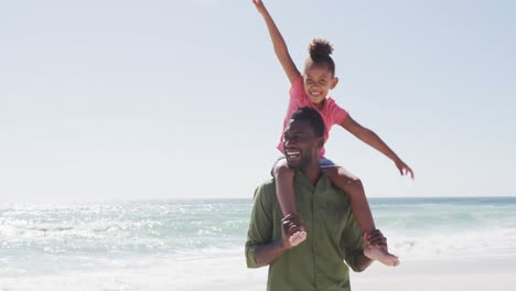 Lächelnder-Afroamerikanischer-Vater,-Der-Seine-Tochter-Am-Sonnigen-Strand-Trägt