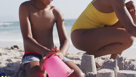 Smiling-african-american-son-with-mother-building-sandcastle-on-sunny-beach
