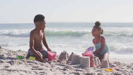 Portrait-of-smiling-african-american-siblings-building-sandcastle-with-american-flag-on-sunny-beach