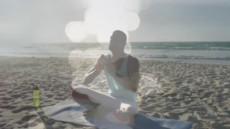 Animation-of-glowing-lights-over-african-american-man-doing-yoga-meditation-on-beach