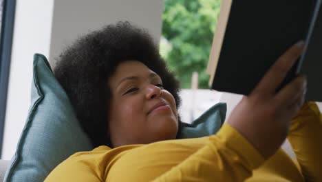 Video-Einer-Glücklichen-Afroamerikanerin-In-Übergröße,-Die-Auf-Dem-Sofa-Liegt-Und-Ein-Buch-Liest