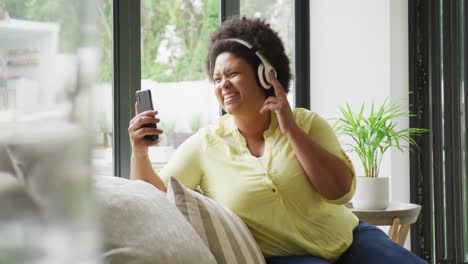 Video-of-happy-plus-size-african-american-woman-with-headphones-on-sofa,-having-video-call