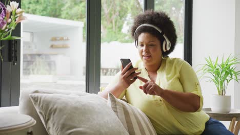 Video-of-happy-plus-size-african-american-woman-with-headphones-sitting-on-sofa-with-smartphone
