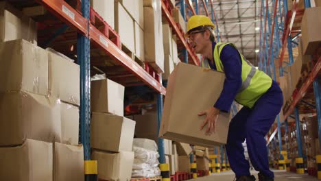 Asian-male-worker-wearing-safety-suit-with-helmet-and-carrying-boxes-in-warehouse