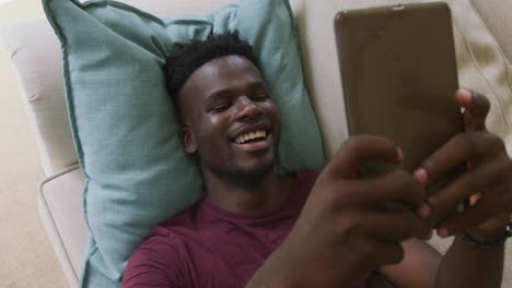 Video-of-happy-african-american-man-lying-on-sofa-and-using-smartphone