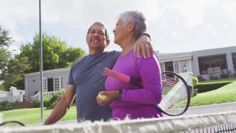 Video-Eines-Glücklichen-Gemischtrassigen-älteren-Paares,-Das-Sich-Umarmt-Und-Mit-Schlägern-Auf-Dem-Tennisplatz-Läuft