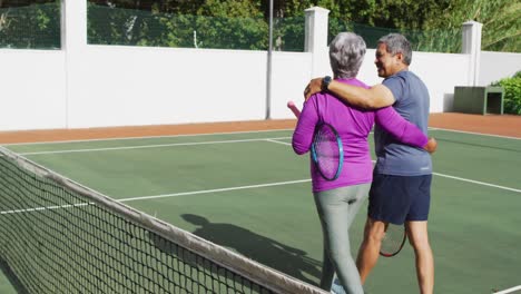 Video-of-happy-biracial-senior-couple-embracing-and-walking-with-rackets-on-tennis-court