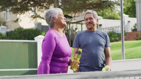Video-Eines-Glücklichen-Gemischtrassigen-Seniorenpaares,-Das-Wasser-Und-Tennisbälle-Auf-Dem-Tennisplatz-Hält