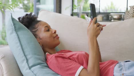 Video-of-happy-african-american-woman-lying-on-sofa-and-using-smartphone