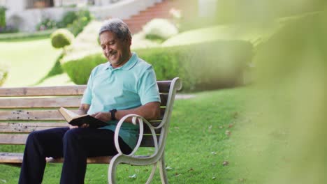 Video-of-happy-biracial-senior-man-reading-book-and-sitting-on-bench-in-garden