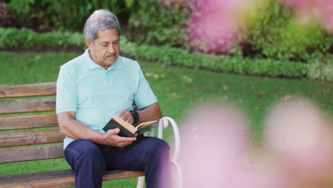 Video-of-relaxed-biracial-senior-man-reading-book-and-sitting-on-bench-in-garden
