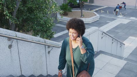 Happy-plus-size-biracial-woman-walking-on-stairs-and-talking-on-smartphone-in-city