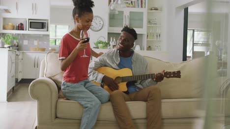 Video-of-happy-african-american-couple-sitting-on-sofa,-singing-and-playing-guitar