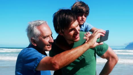 Animation-of-diverse-icons-over-caucasian-grandfather,-father-and-son-taking-selfie-on-beach