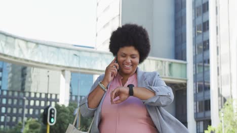 Happy-plus-size-biracial-woman-checking-smartwatch-and-walking-in-city