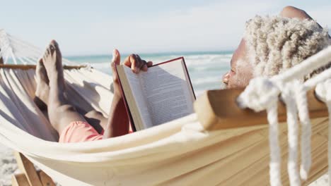 Senior-african-american-man-reading-and-lying-in-hammock-on-sunny-beach
