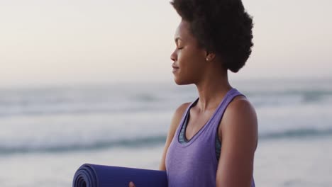 Portrait-of-happy-african-american-woman-with-yoga-mat-on-sunny-beach